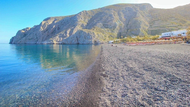 A pebble beach with a mountain backdrop, clear water, and a building with outdoor seating. - Sail Inn (Santorini)