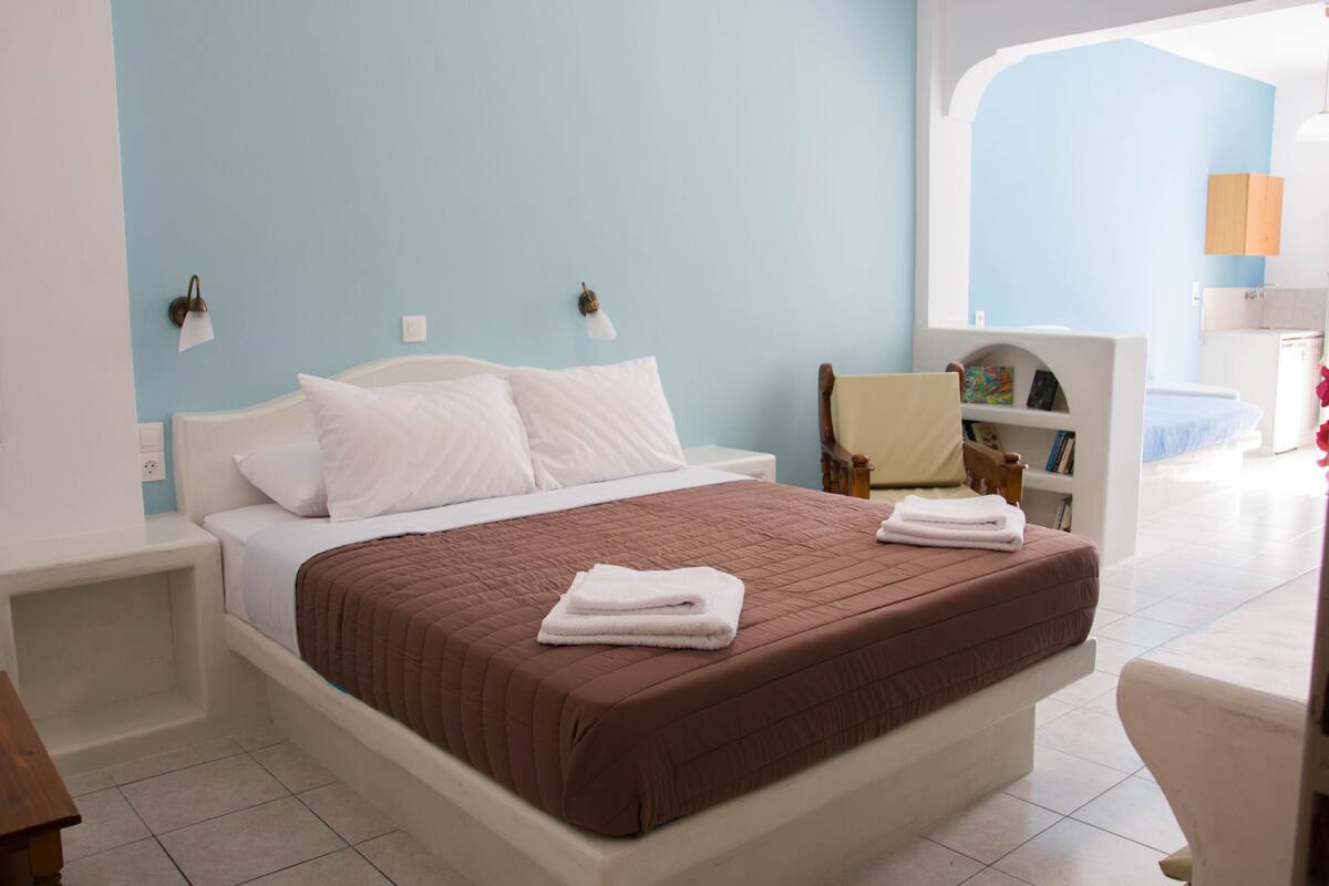 Una cama bien hecha con cubrecama marrón y almohadas blancas, una mesita de noche y una silla con una estantería.