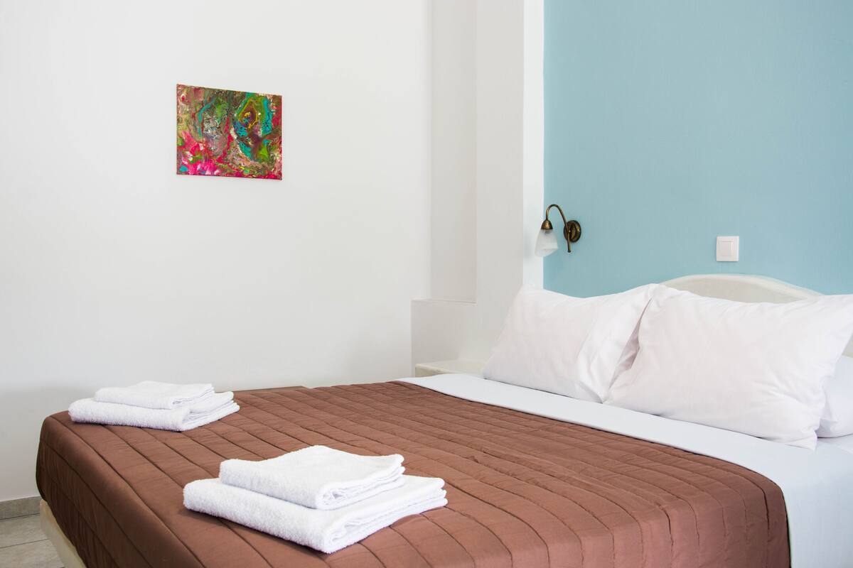 Una habitación de hotel con una colcha marrón, almohadas blancas, un pequeño cuadro abstracto en la pared y una lámpara de noche.