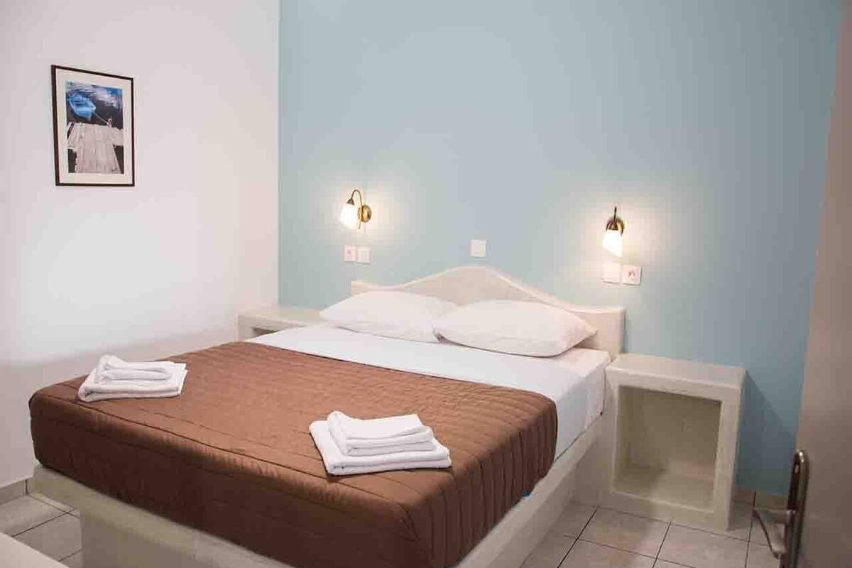 Una cama bien hecha con sábanas blancas y una colcha marrón, dos mesitas de noche y lámparas fijadas a la pared.