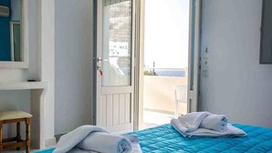 A hotel room with a bed, a chair, a door, and a view of the sea. - Sail Inn (Santorini)