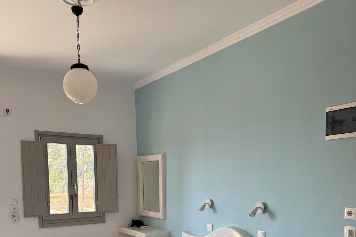 Una cama bien hecha con un edredón marrón, una almohada blanca y dos toallas dobladas. Hay una mesita de noche con una lámpara, un espejo en la pared y una ventana con persianas cerradas.