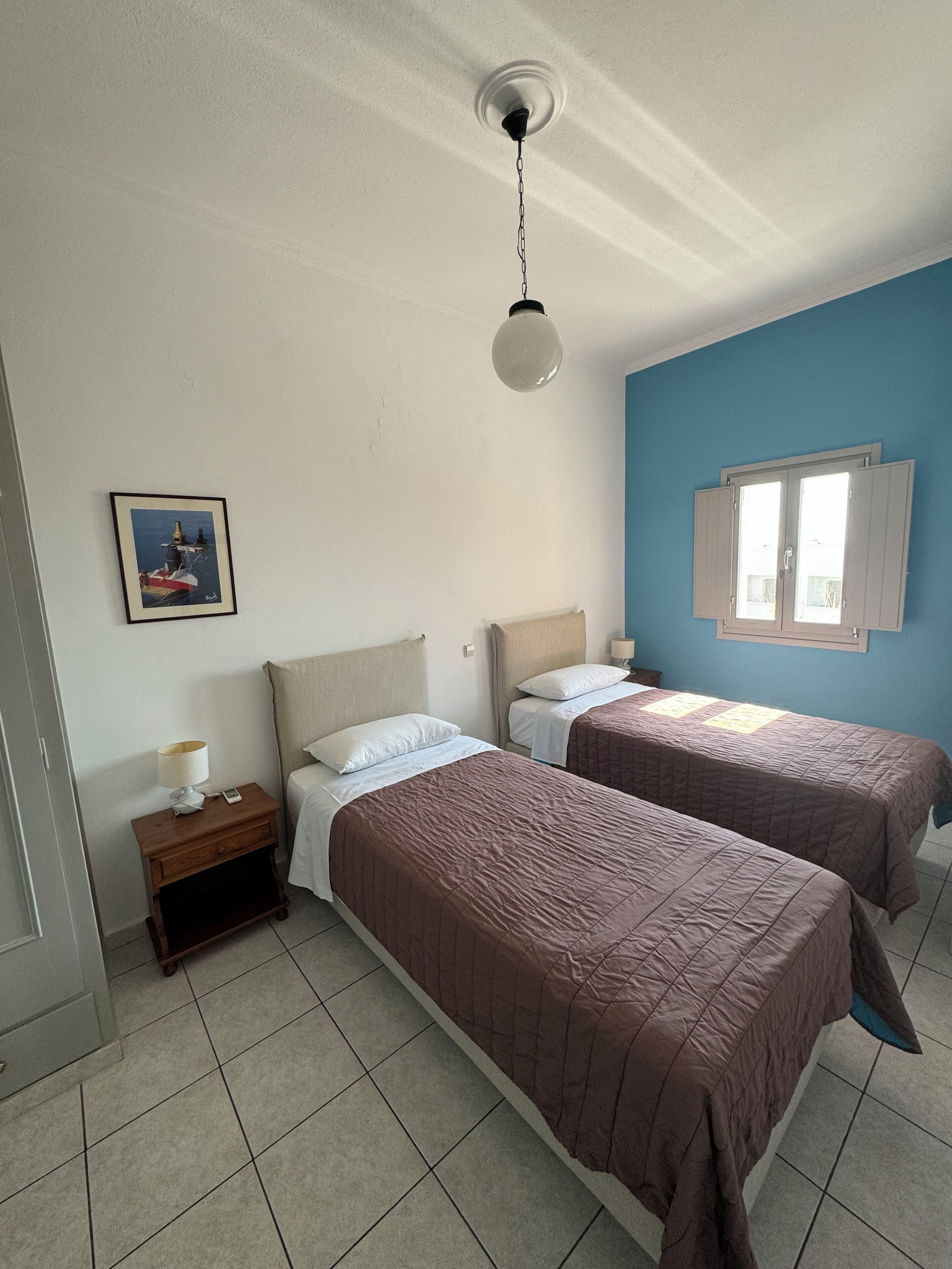 Une chambre avec deux lits, une table de chevet, une lampe, un tableau et une fenêtre avec des volets.