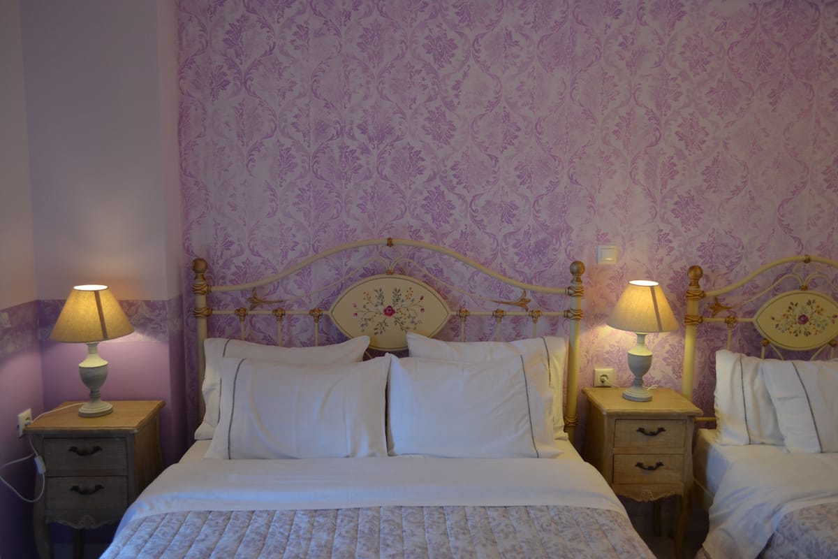 Un dormitorio con dos camas, cada una con cabecero de madera y una mesita de noche con lámpara. Las paredes tienen un estampado de papel tapiz floral.