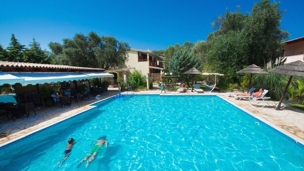 Una piscina con agua cristalina azul, rodeada de sillas de descanso y sombrillas, y un edificio con tejado de baldosas al fondo.