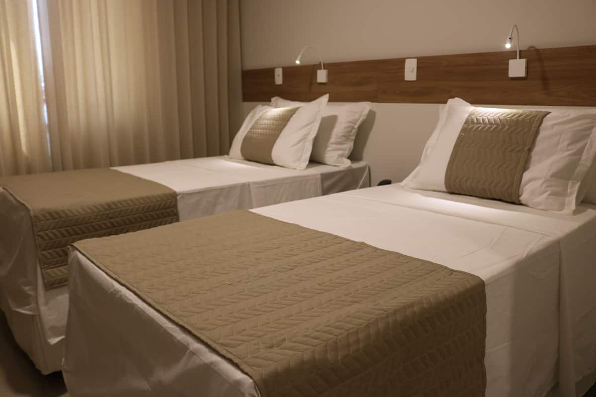 A hotel room with two single beds, each with a beige quilted bedspread and white pillows. There are two bedside tables with lamps, and the room has a wooden headboard with wall-mounted light fixtures.