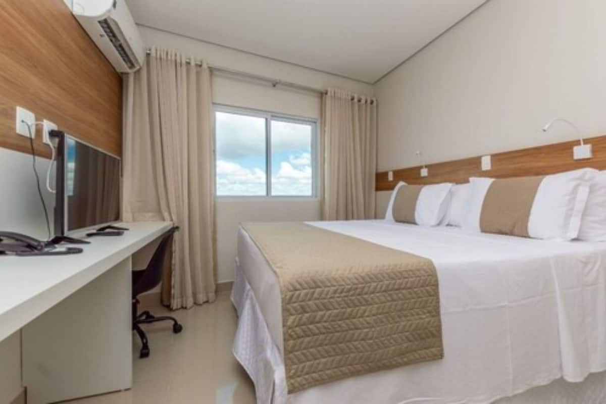 A hotel room with a bed, a desk with a computer, and a window with curtains.