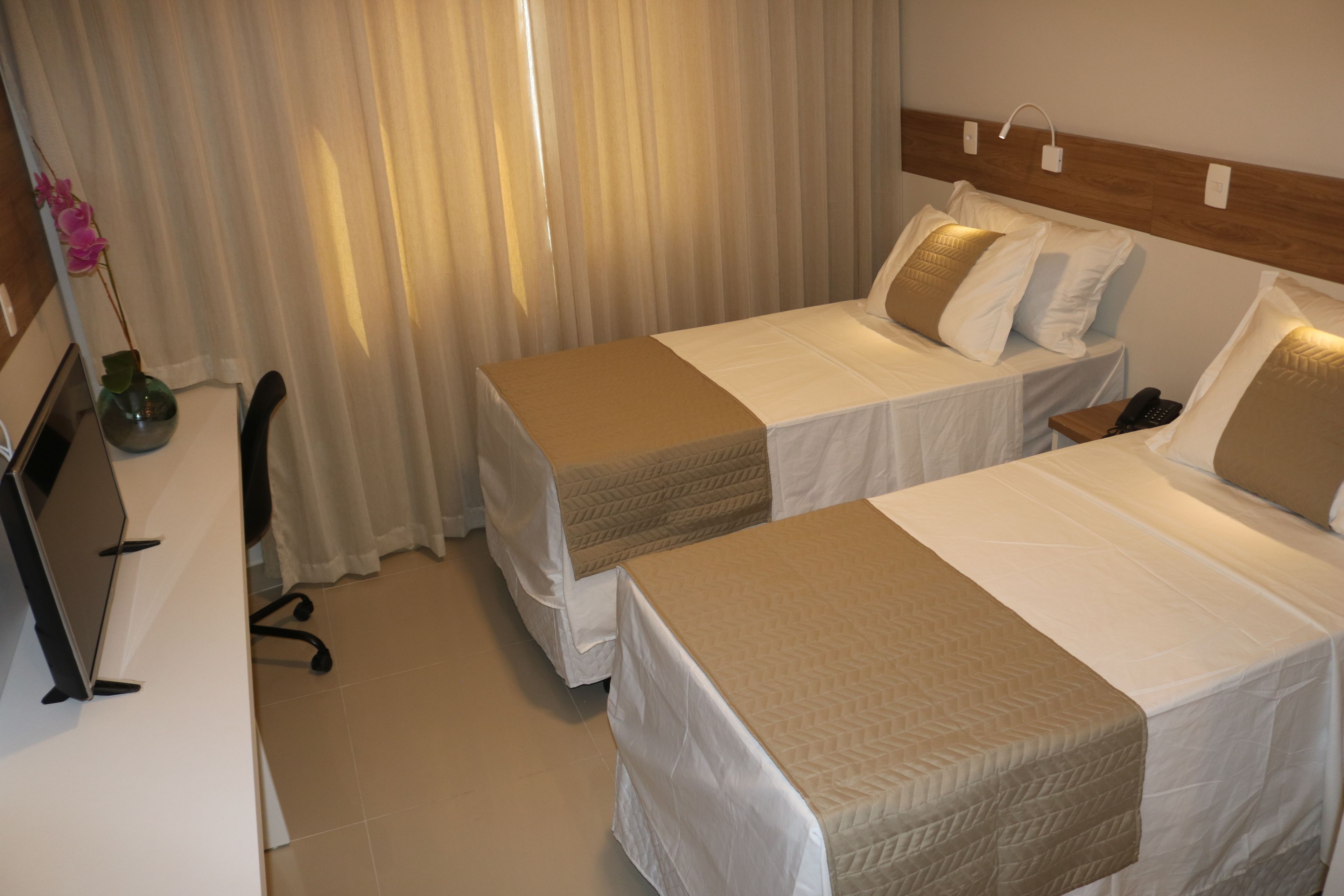 Two single beds with beige bedspreads, a desk with a chair, and a vase with flowers.