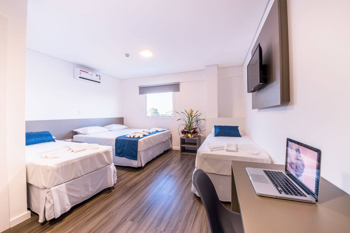 A hotel room with two single beds, a desk with a laptop, a TV on the wall, and a window with a view.