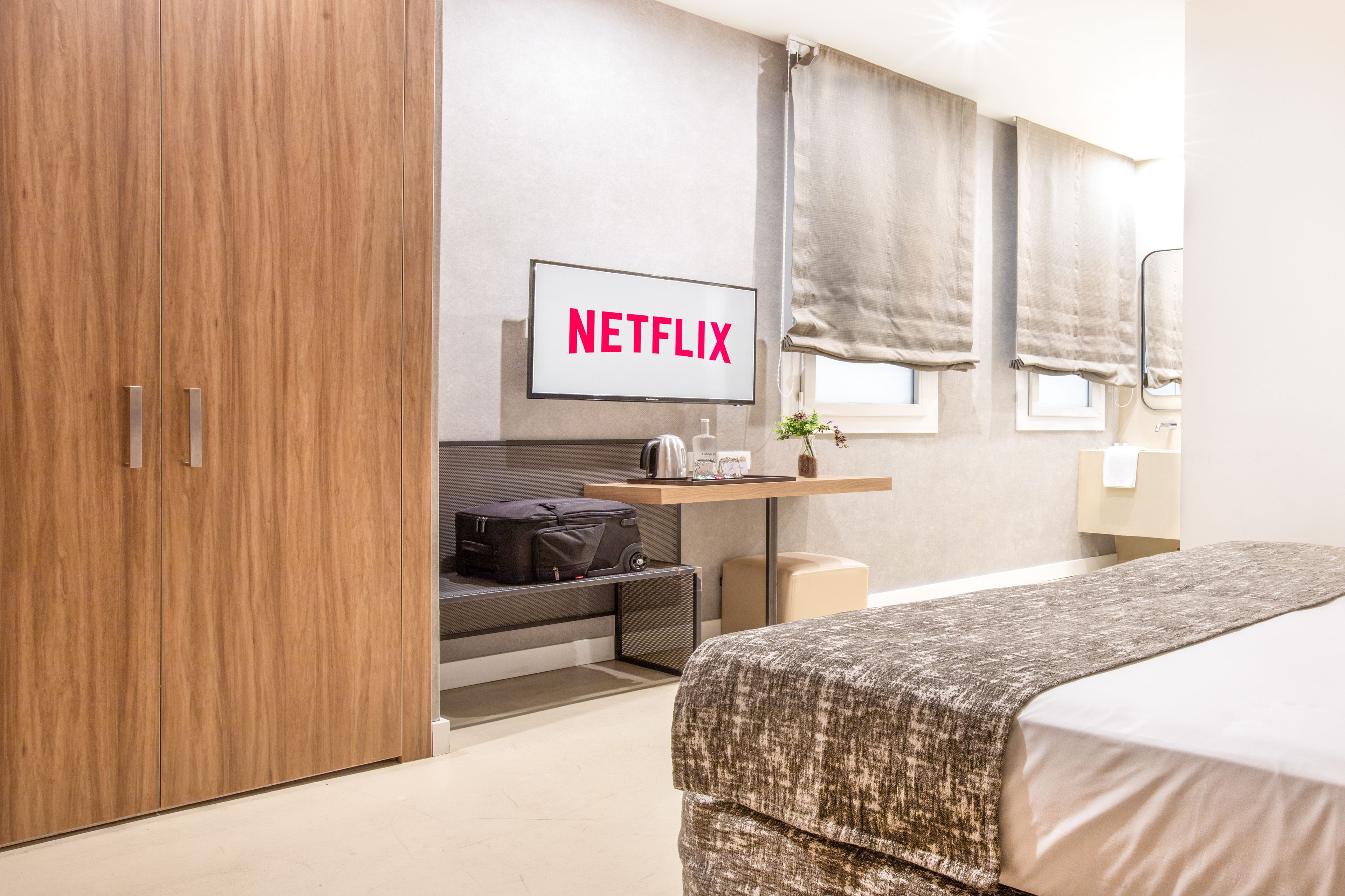 Une chambre d’hôtel moderne avec une armoire en bois, un lit recouvert d’une couverture à motifs, une petite table avec une bouilloire et un panneau mural indiquant « NETFLIX ».