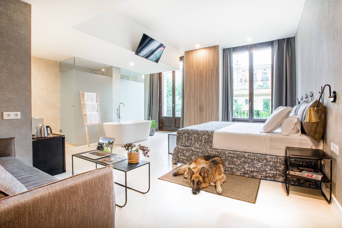 Ein modernes Hotelzimmer mit einer Couch, einem Bett, einem Laptop, einem Hund, einem Fernseher, einer Badewanne und einem Fenster mit Blick auf Grünflächen.