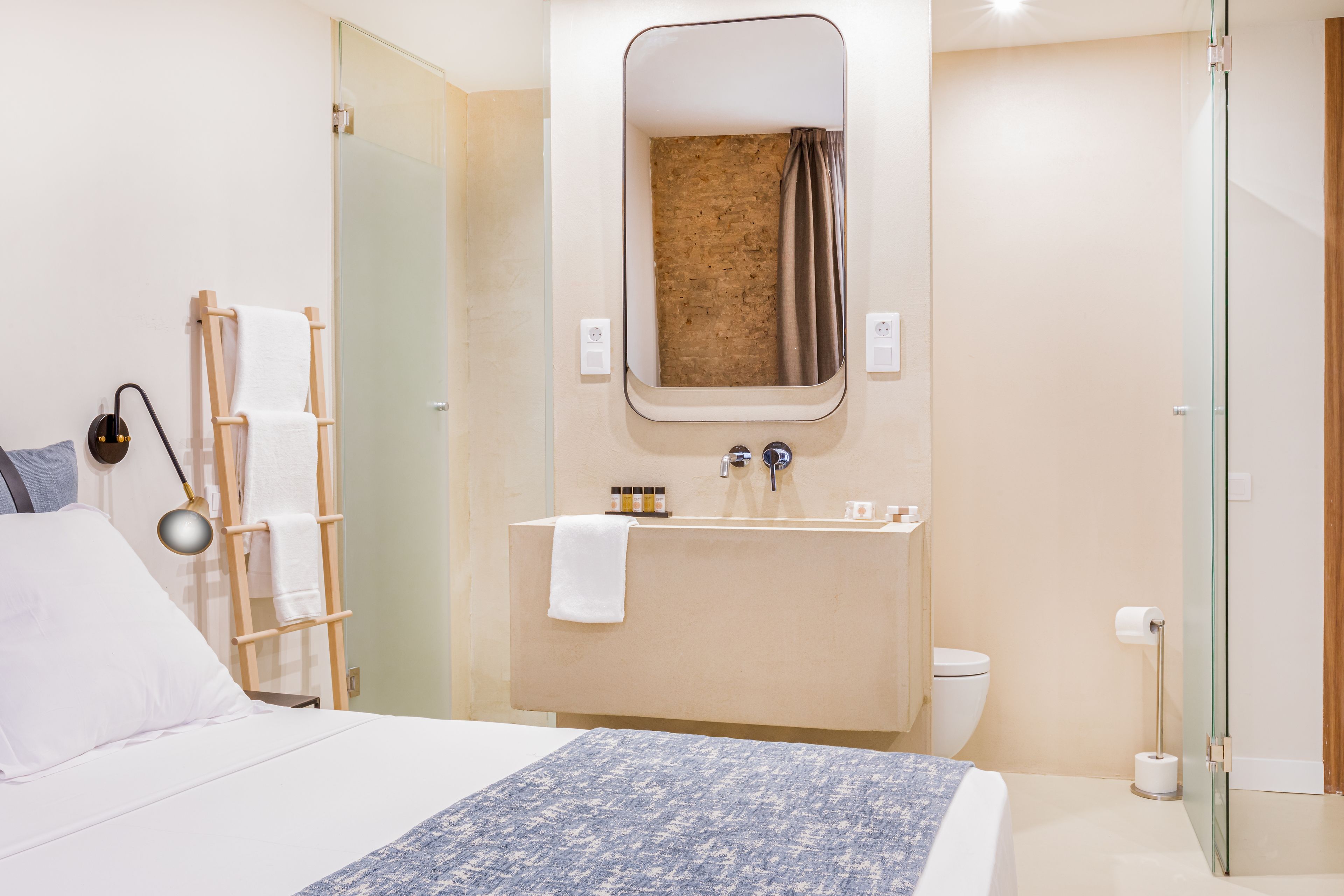 A hotel room with a bed, a bedside table, a mirror, a sink, and a toilet.