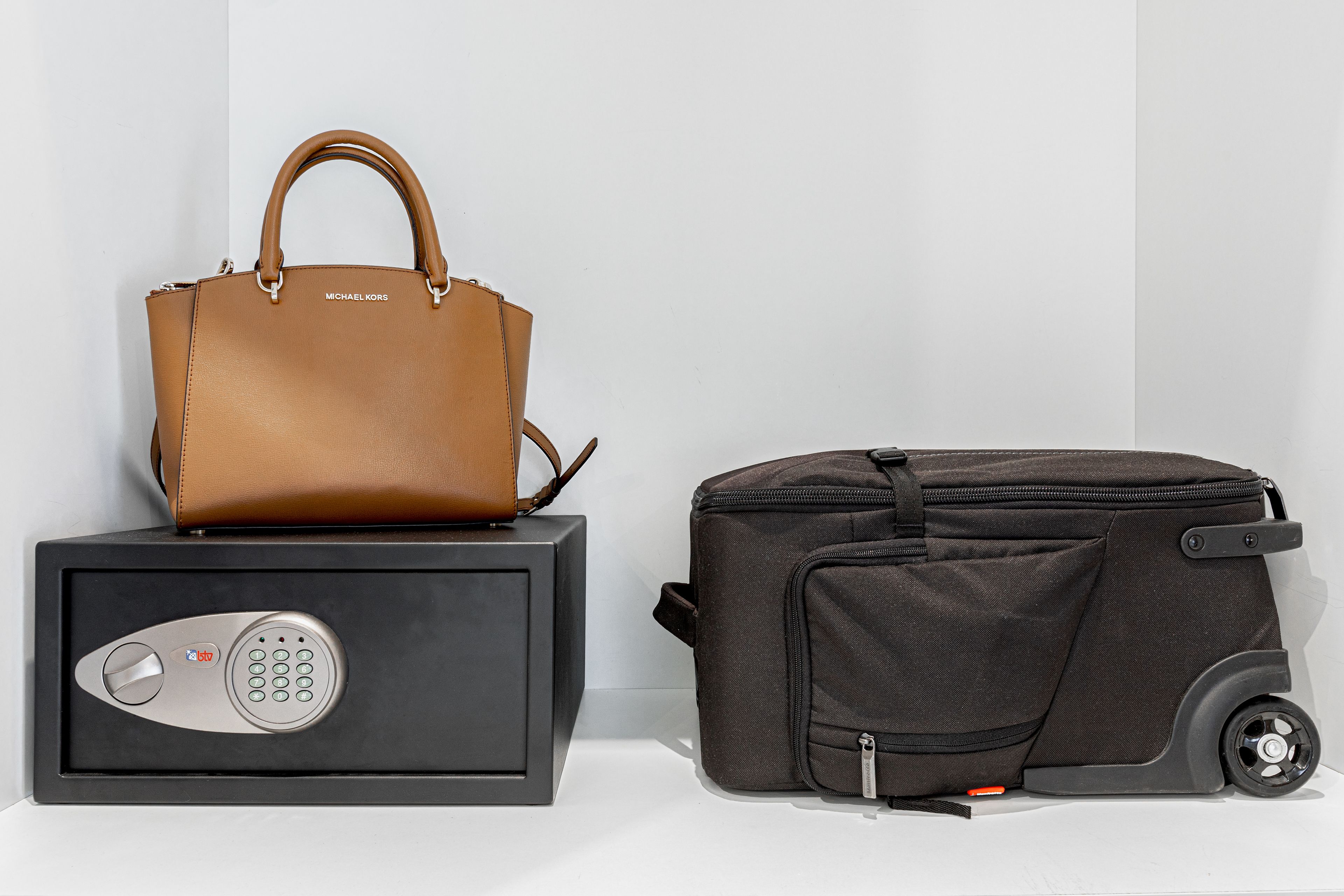 A brown leather handbag and a black wheeled suitcase next to a black safe with a keypad lock.