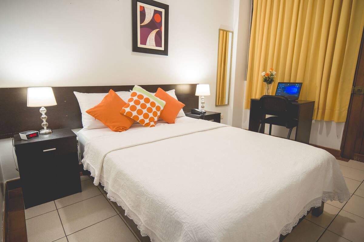 A neatly made bed with white and orange bedding, a digital clock on a bedside table, and a laptop on a desk with a vase of flowers.