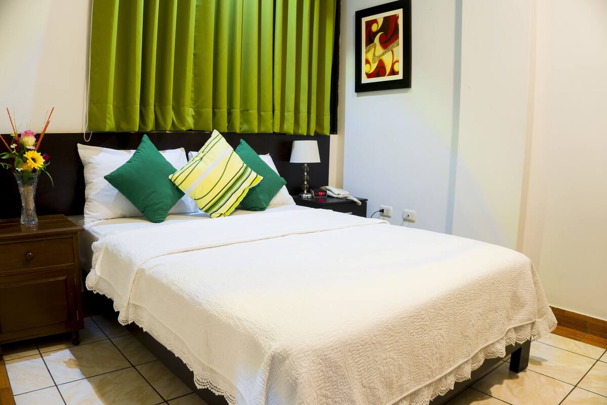 A neatly made bed with white linens and green and yellow striped pillows, a bedside lamp, and a vase with flowers on a wooden nightstand.