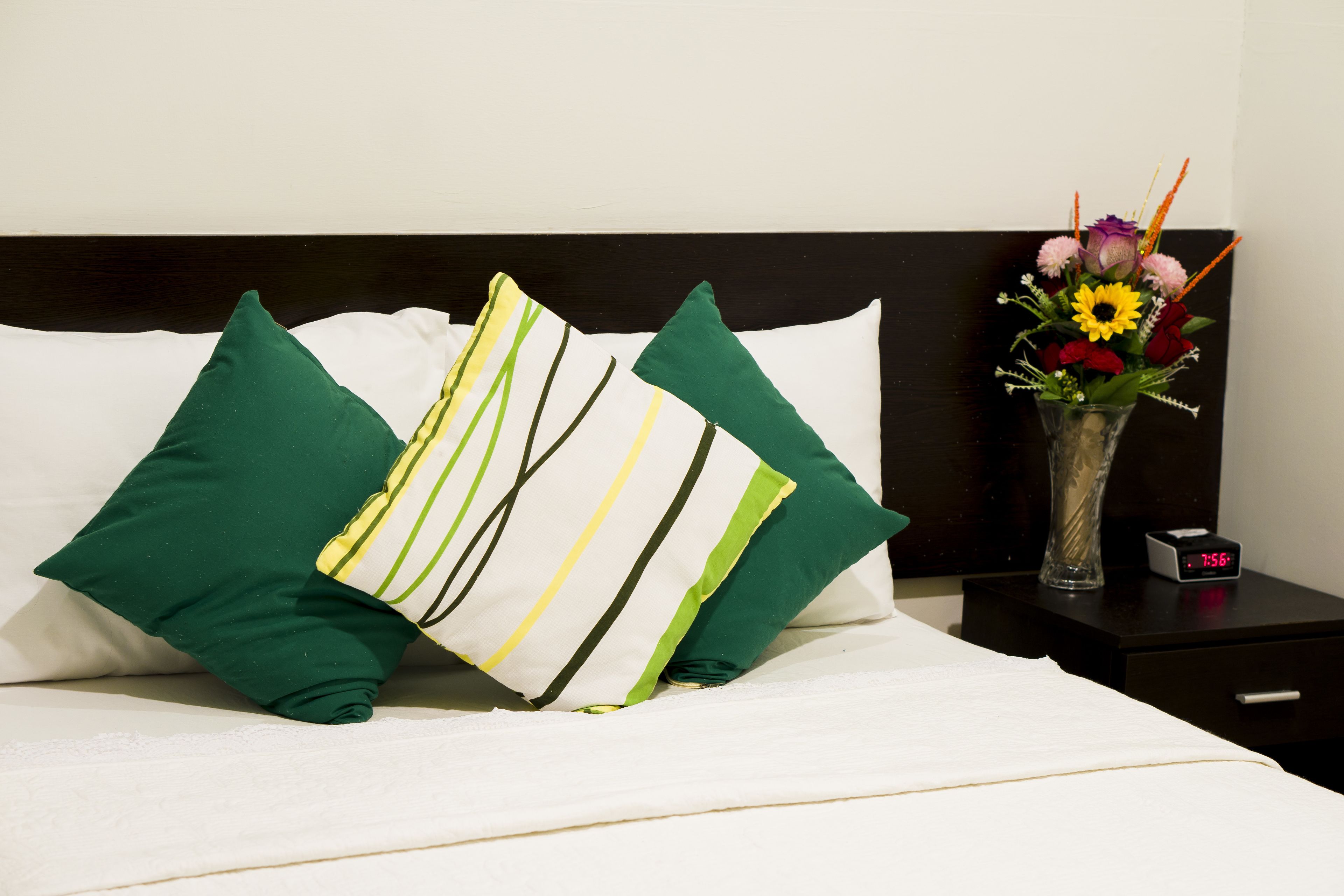 A neatly made bed with green and white striped pillows, a vase with flowers, and a digital clock on a bedside table.