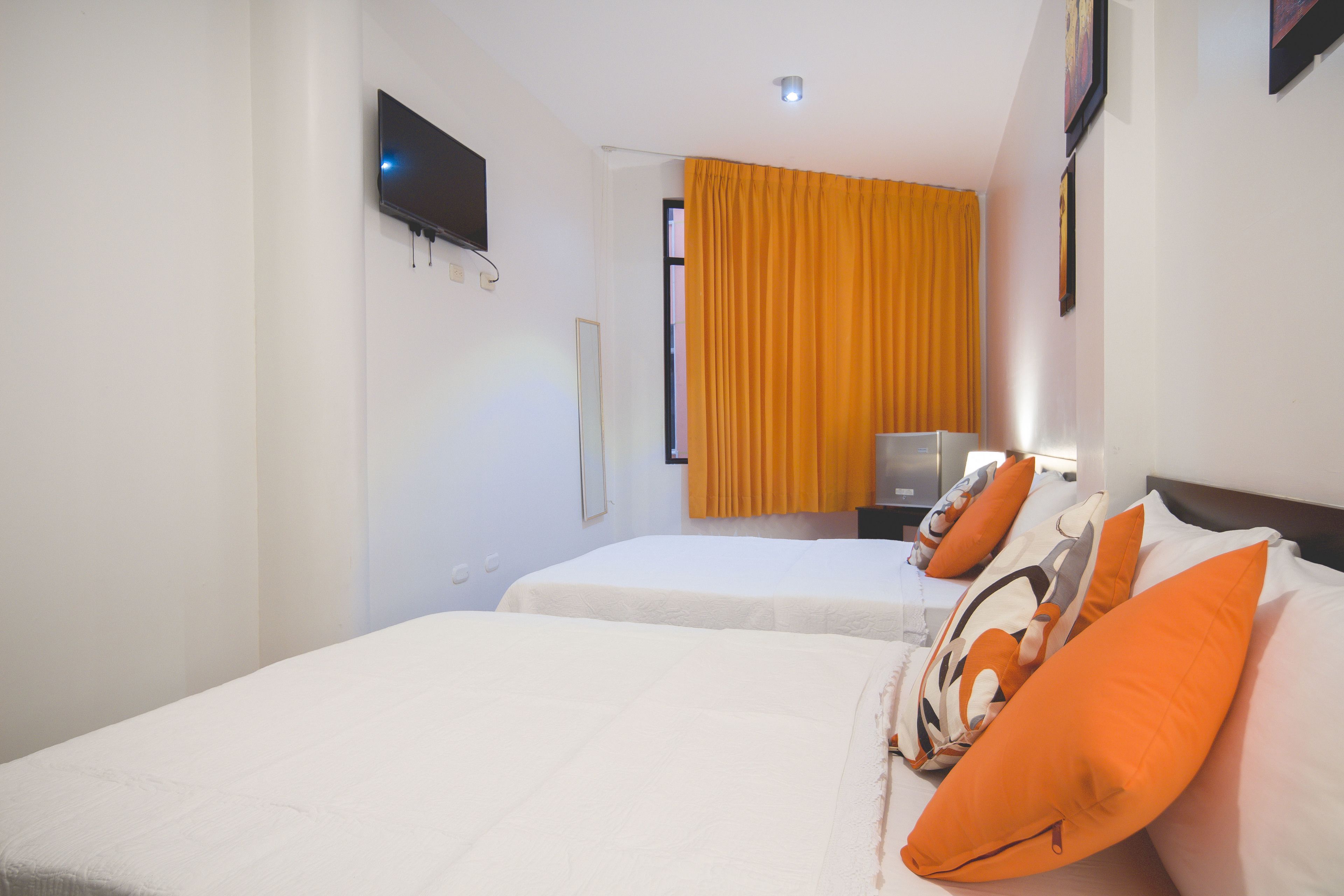 A hotel room with two beds, a television mounted on the wall, and a window with yellow curtains.