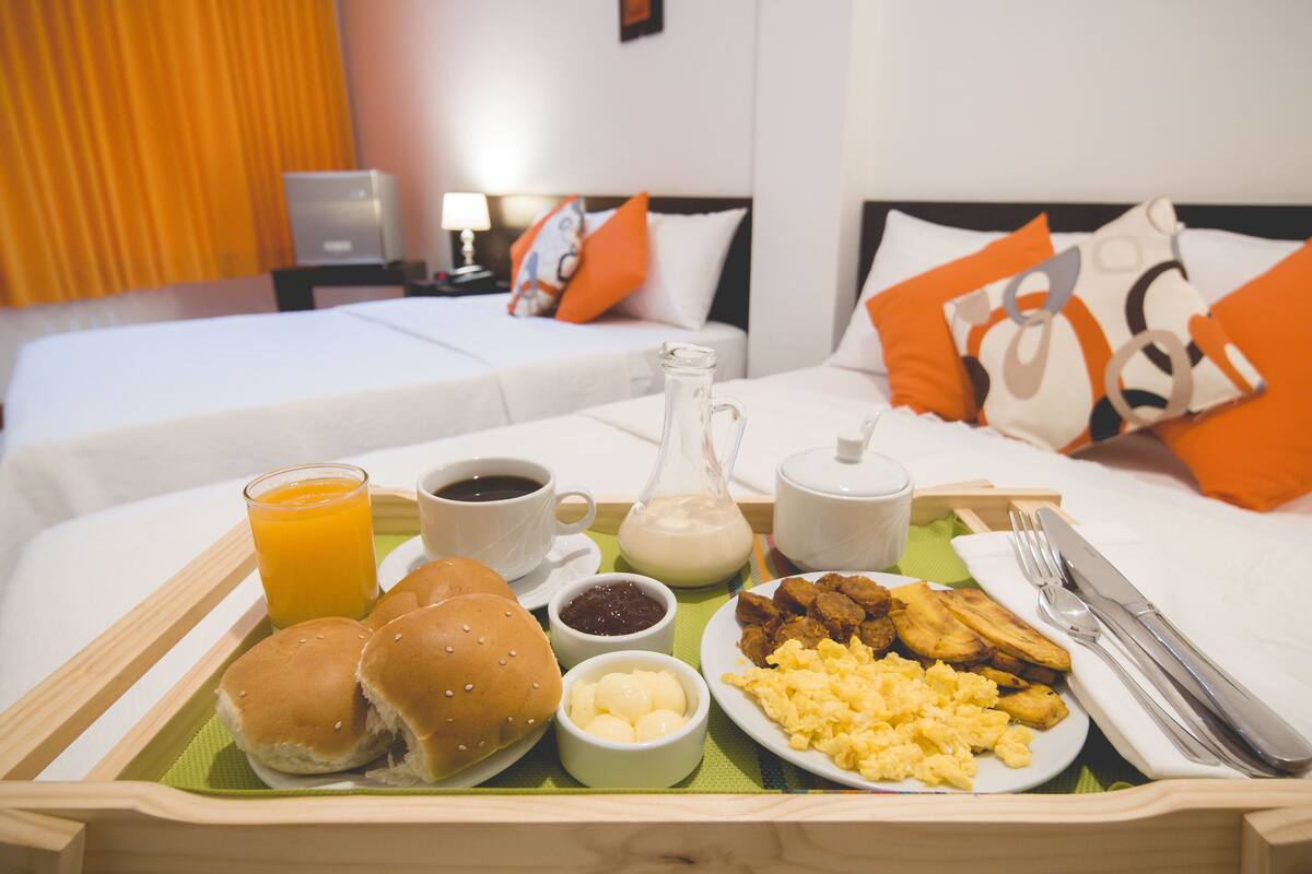 A breakfast tray with eggs, sausage, toast, and juice on a bed with two pillows and a nightstand with a lamp.