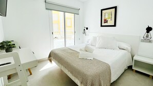 A bedroom with a bed, a desk, a chair, a plant, and a sliding door to a balcony.