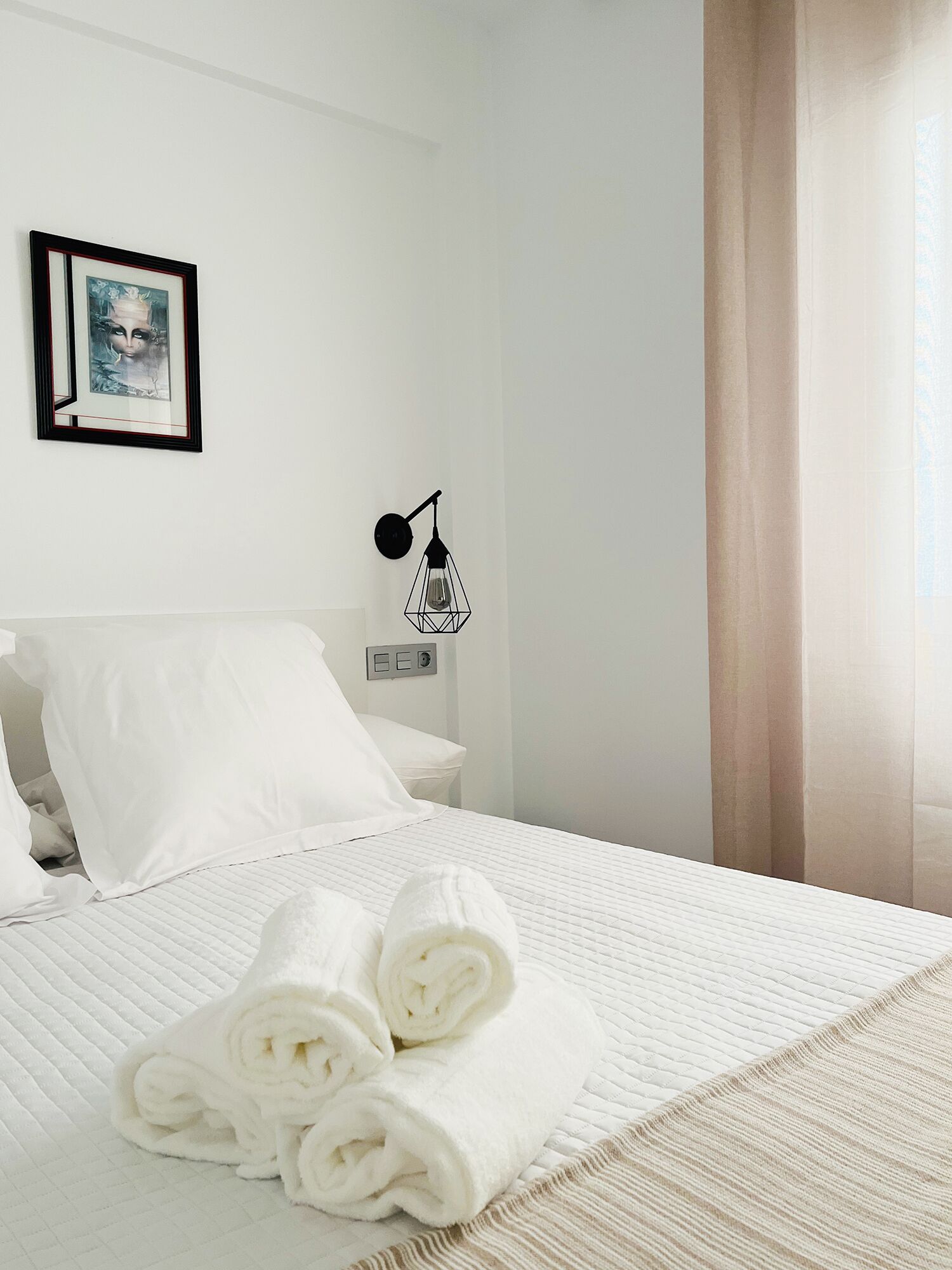Een net opgemaakt bed met opgerolde handdoeken, een ingelijst beeld aan de muur, een wandlamp en een raam met gordijnen.