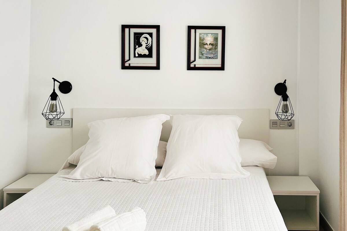 Quarto de hotel com uma cama, duas mesas de cabeceira e duas luminárias fixadas na parede.