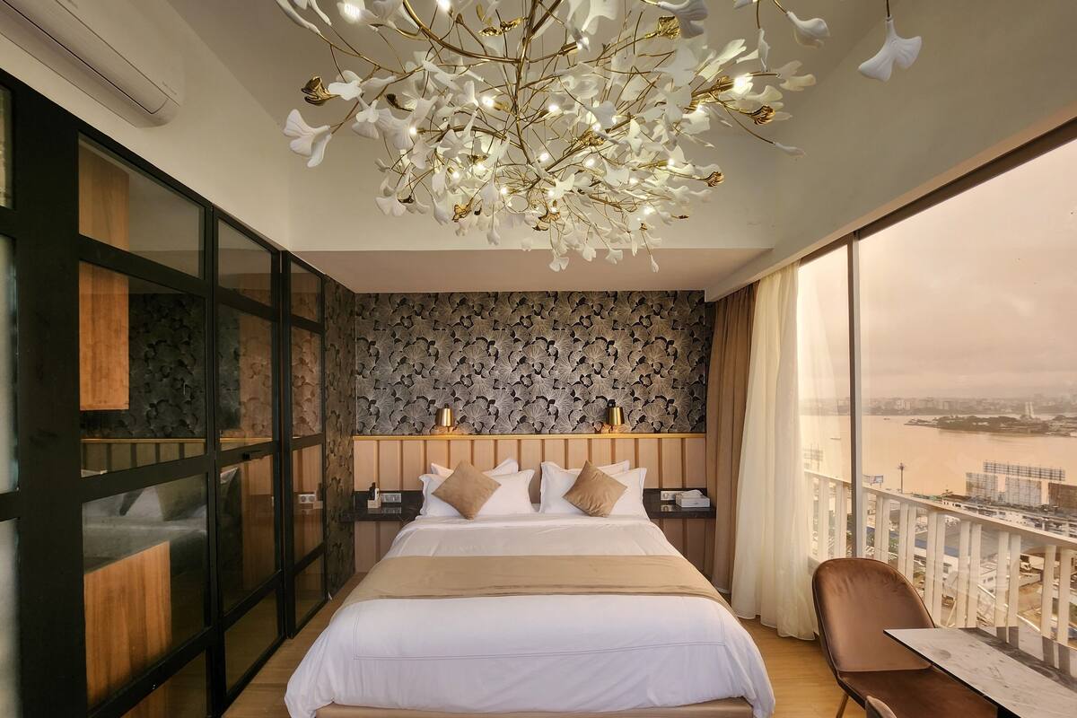 A modern hotel room with a large bed, a view of a river, and a balcony with a table and chair.