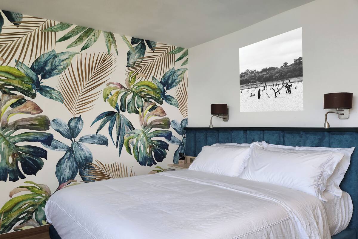 A bedroom with a large bed, a blue headboard, a tropical leaf wallpaper, two wall-mounted lamps, and a framed picture on the wall.