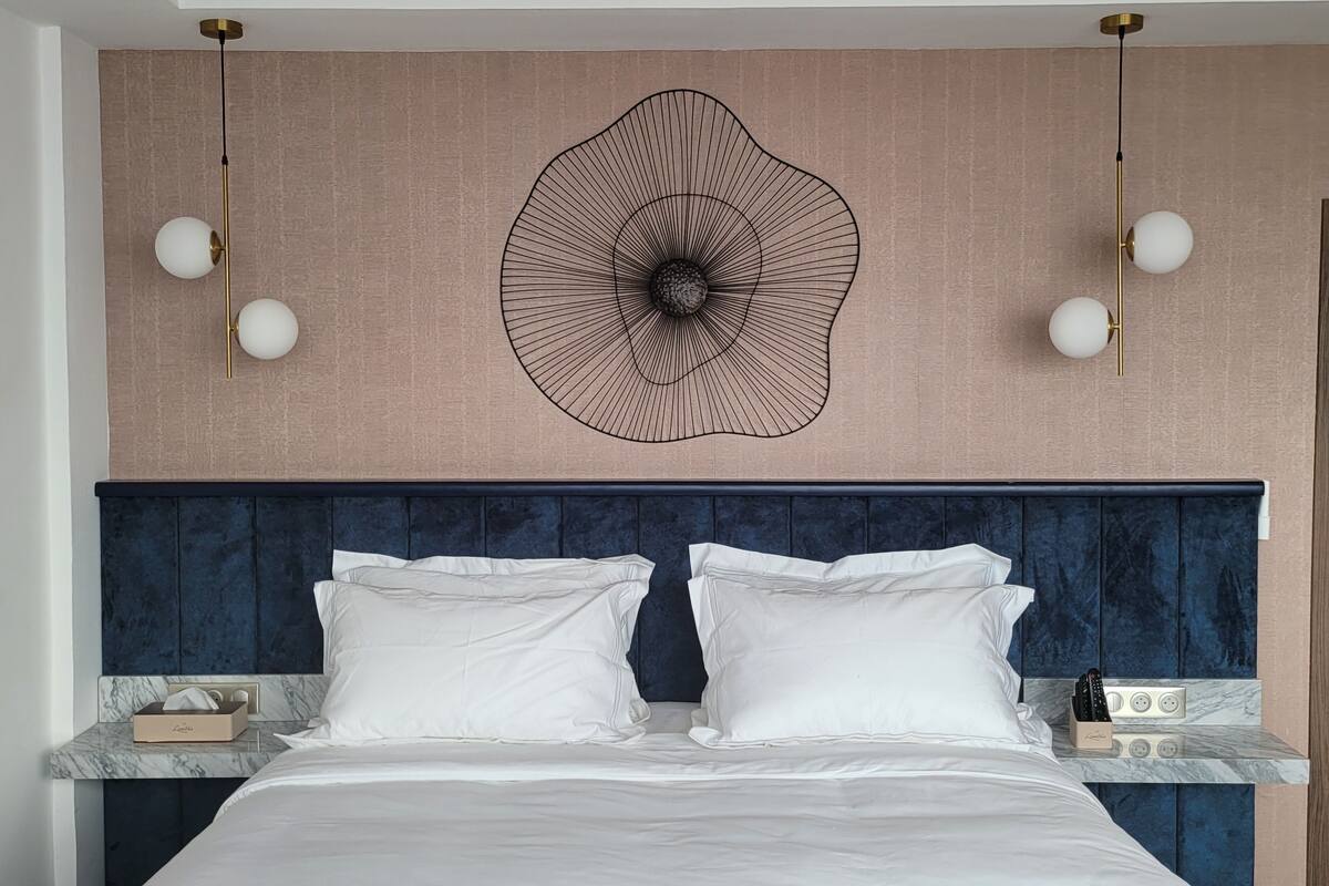 A modern hotel room with a large bed, a dark blue headboard, and a unique wall art piece above the bed.