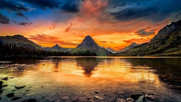 A mountain lake with clear reflections, surrounded by forested mountains and a dramatic sunset sky.