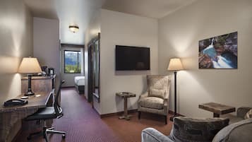 Habitación de hotel con escritorio, silla de oficina, sillón y una mesita auxiliar. Hay un televisor montado en la pared y un cuadro encima.