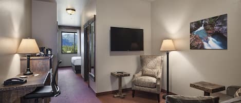 A hotel room with a desk, office chair, armchair, and a small side table. There is a TV mounted on the wall and a painting above it.