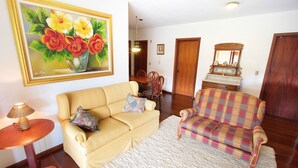 A living room with a yellow sofa, a plaid armchair, a small round table with a lamp, and a painting of flowers on the wall. - My Home - Titisee (Gramado)