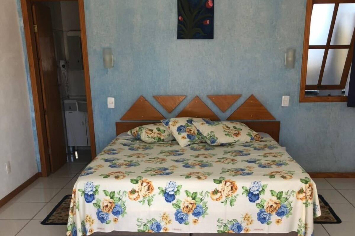 A bedroom with a bed, floral bedspread, a painting, an air conditioner, and a window.