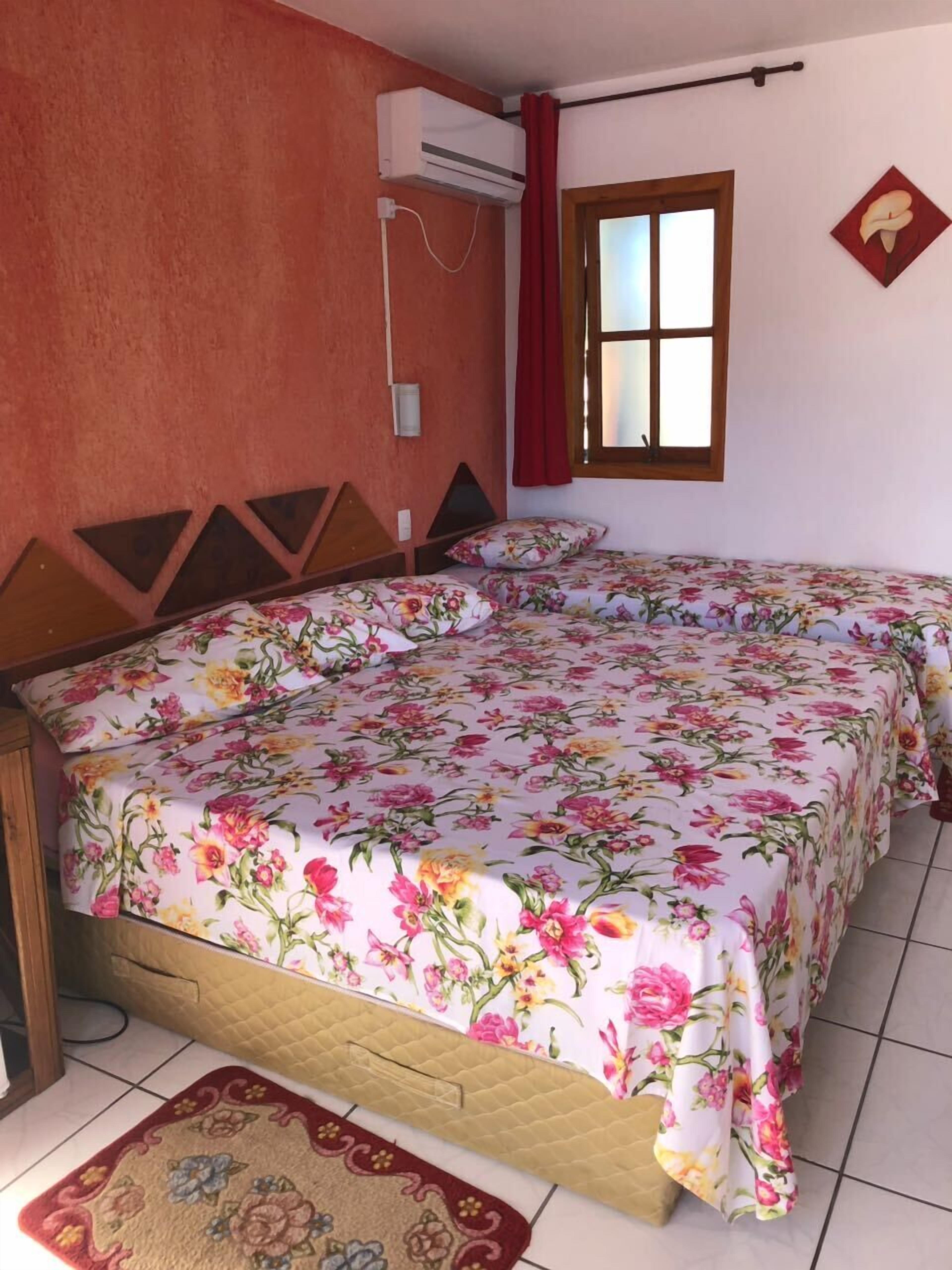 Une chambre avec deux lits, une fenêtre avec des rideaux rouges, un appareil de climatisation et une couvre-lit à motifs floraux.