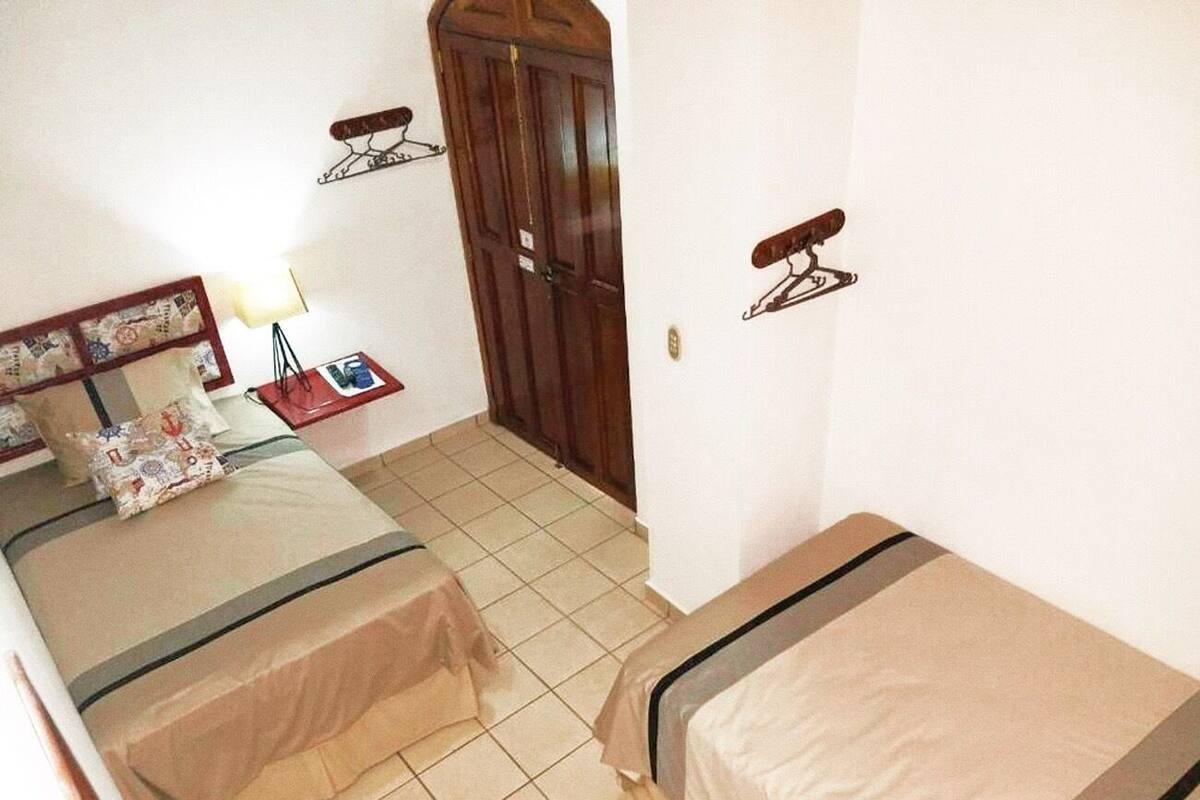 A small, compact room with two beds, a wooden wardrobe, a small table with a lamp, and a wall-mounted shelf.