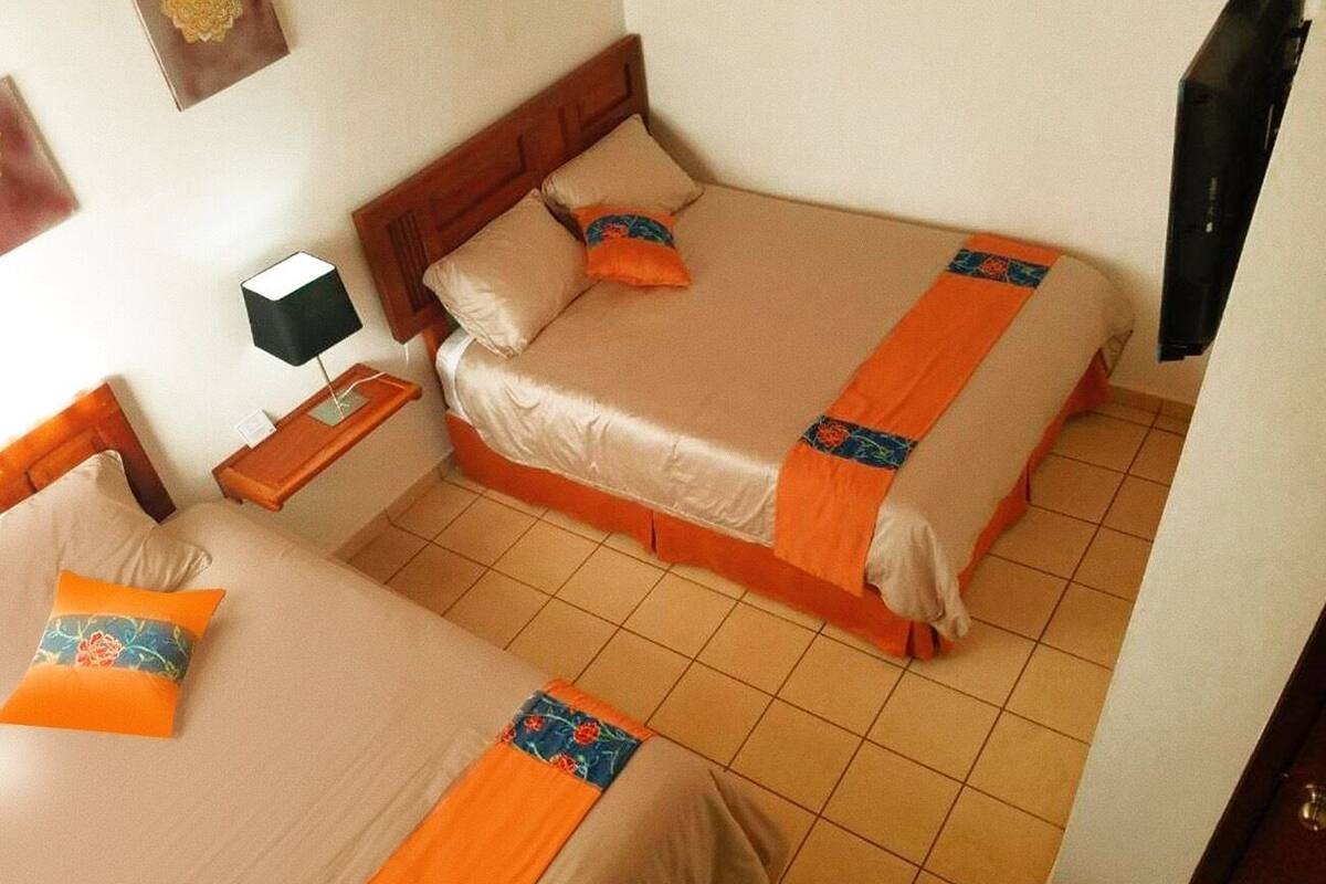 A room with two beds, each with orange and blue bedding, a nightstand with a lamp, and a TV mounted on the wall.