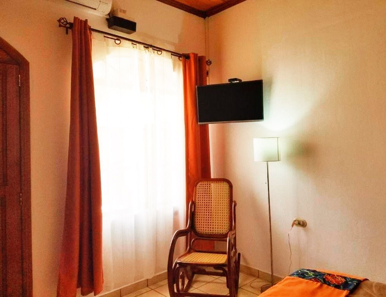 A room with a wooden chair, a television mounted on the wall, and a curtain.