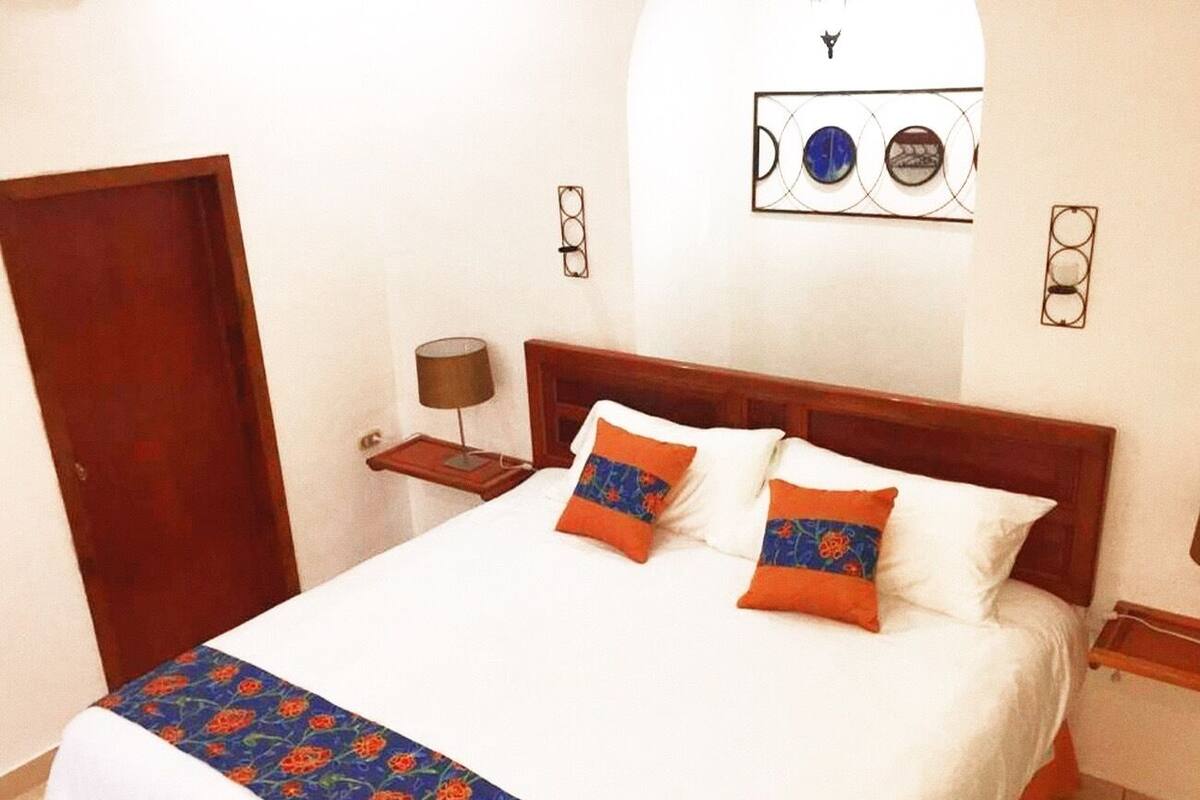 A neatly made bed with a patterned bedspread and two decorative pillows, a wooden headboard, a bedside table with a lamp, and a door to the left.