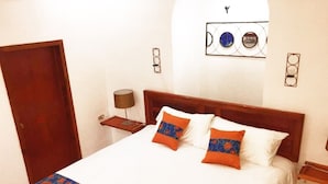 Una cama bien hecha con una colcha estampada y dos almohadas decorativas, un cabecero de madera, una mesita de noche con una lámpara y una puerta a la izquierda.