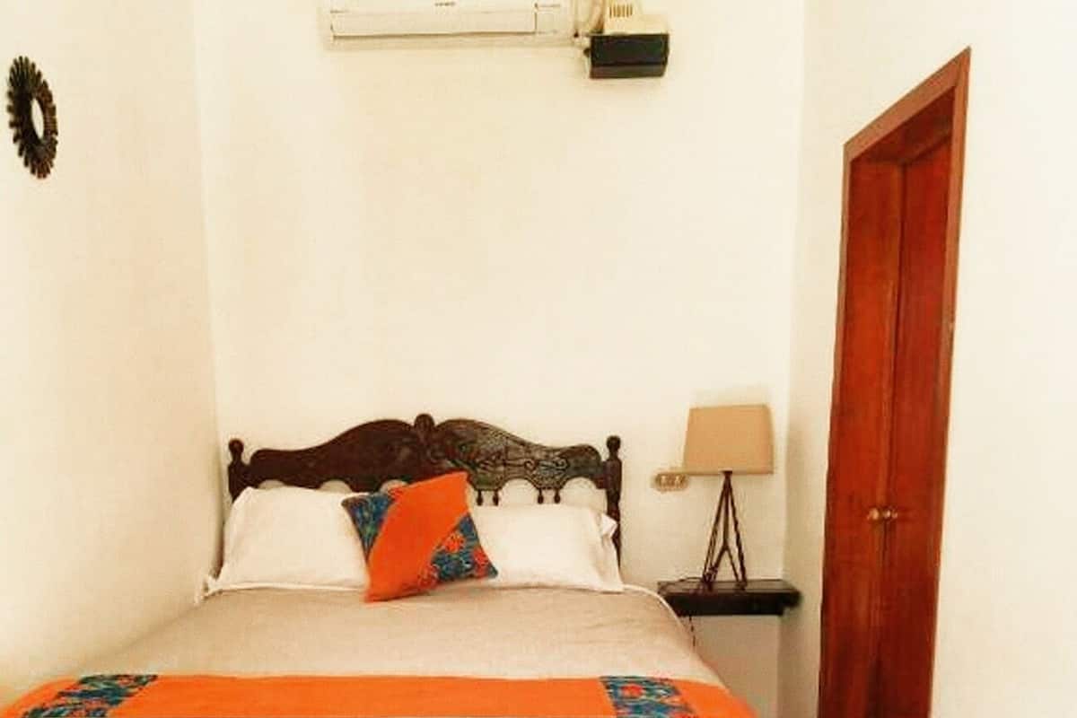 A bedroom with a wooden bed, white bedding, a colorful throw blanket, a bedside lamp, and a wooden door.
