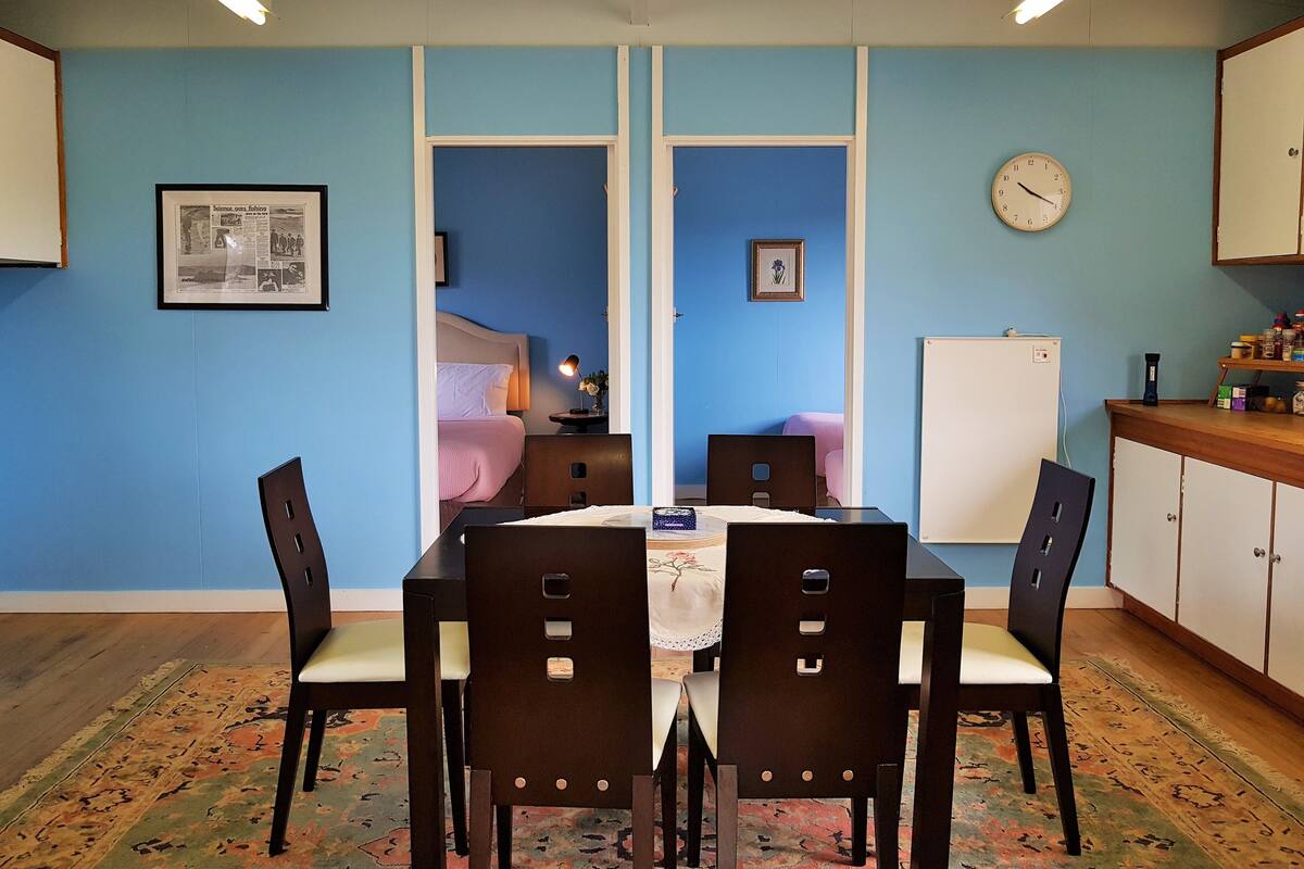 A dining area with a table and chairs, a clock on the wall, and a bed in the background.