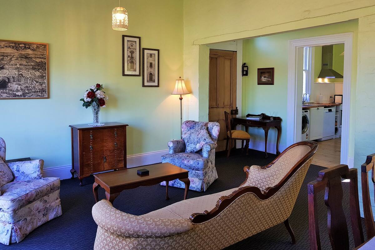 A living room with floral-patterned sofas, a wooden coffee table, and a dining table with chairs. There is a lamp, framed pictures on the wall, and a view into the kitchen through an open doorway.