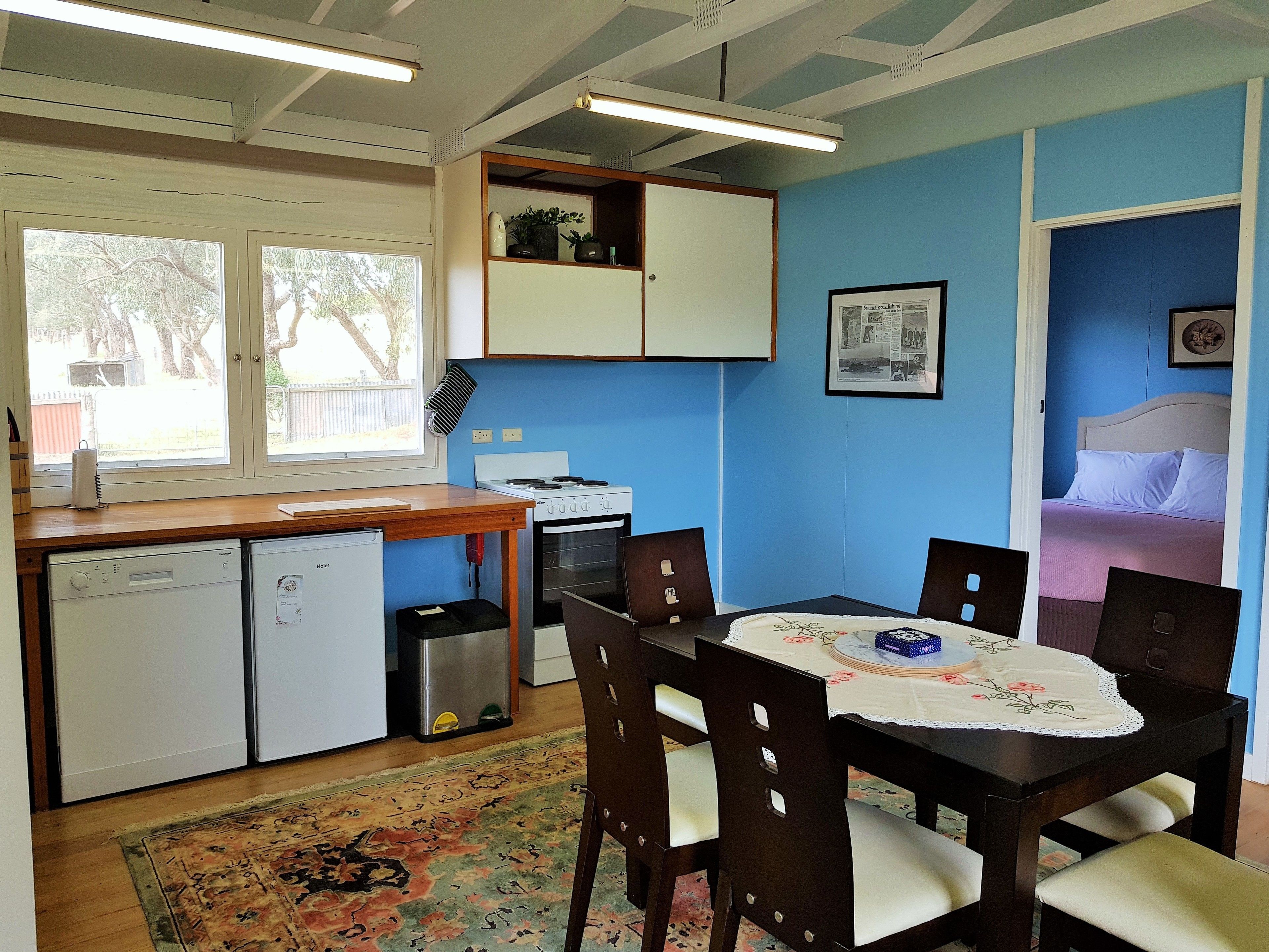 A compact kitchen with a stove, dishwasher, and a small trash bin. There is a dining table with chairs and a rug on the floor.