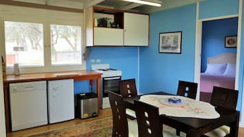 A compact kitchen with a stove, dishwasher, and a small trash bin. There is a dining table with chairs and a rug on the floor.