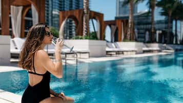Una mujer con traje de baño negro sentada junto a una piscina, con sillas de descanso y palmeras al fondo.