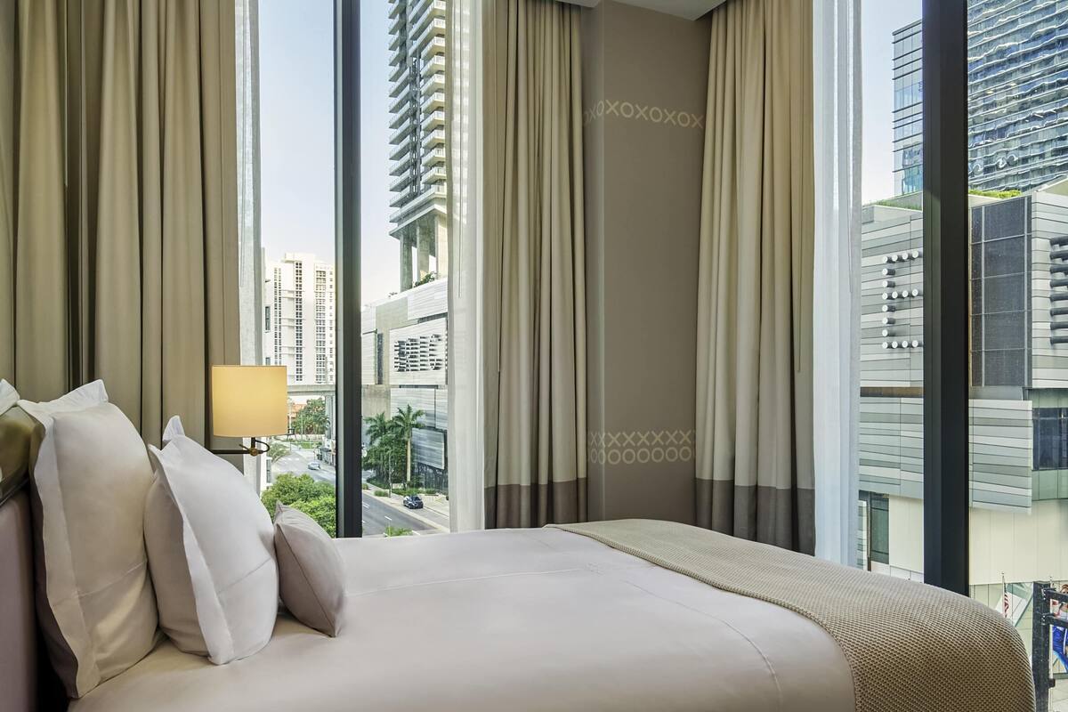 Ein Hotelzimmer mit einem großen Bett, beigen Vorhängen und Blick auf hohe Gebäude.