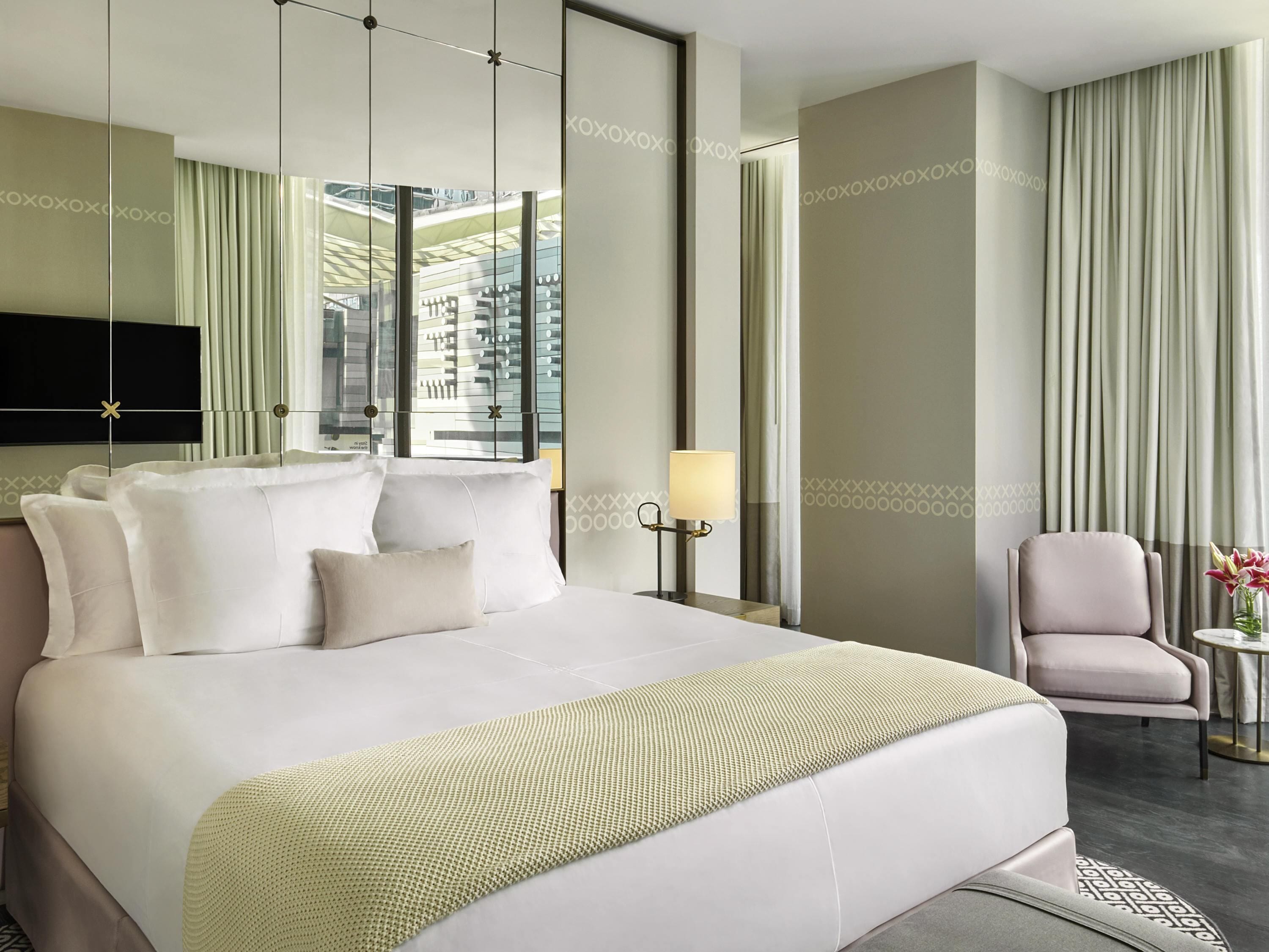 Una habitación de hotel moderna con una cama grande, un televisor de pantalla plana montado en la pared, una mesita de noche con lámpara y vistas a la ciudad a través de ventanales del suelo al techo.