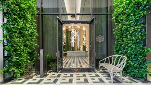 L’entrée d’un hôtel, avec un sol orné de motifs géométriques, un banc et, de part et d’autre, une végétation luxuriante.