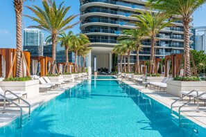 A modern hotel with a central swimming pool, palm trees, and a view of the city skyline.