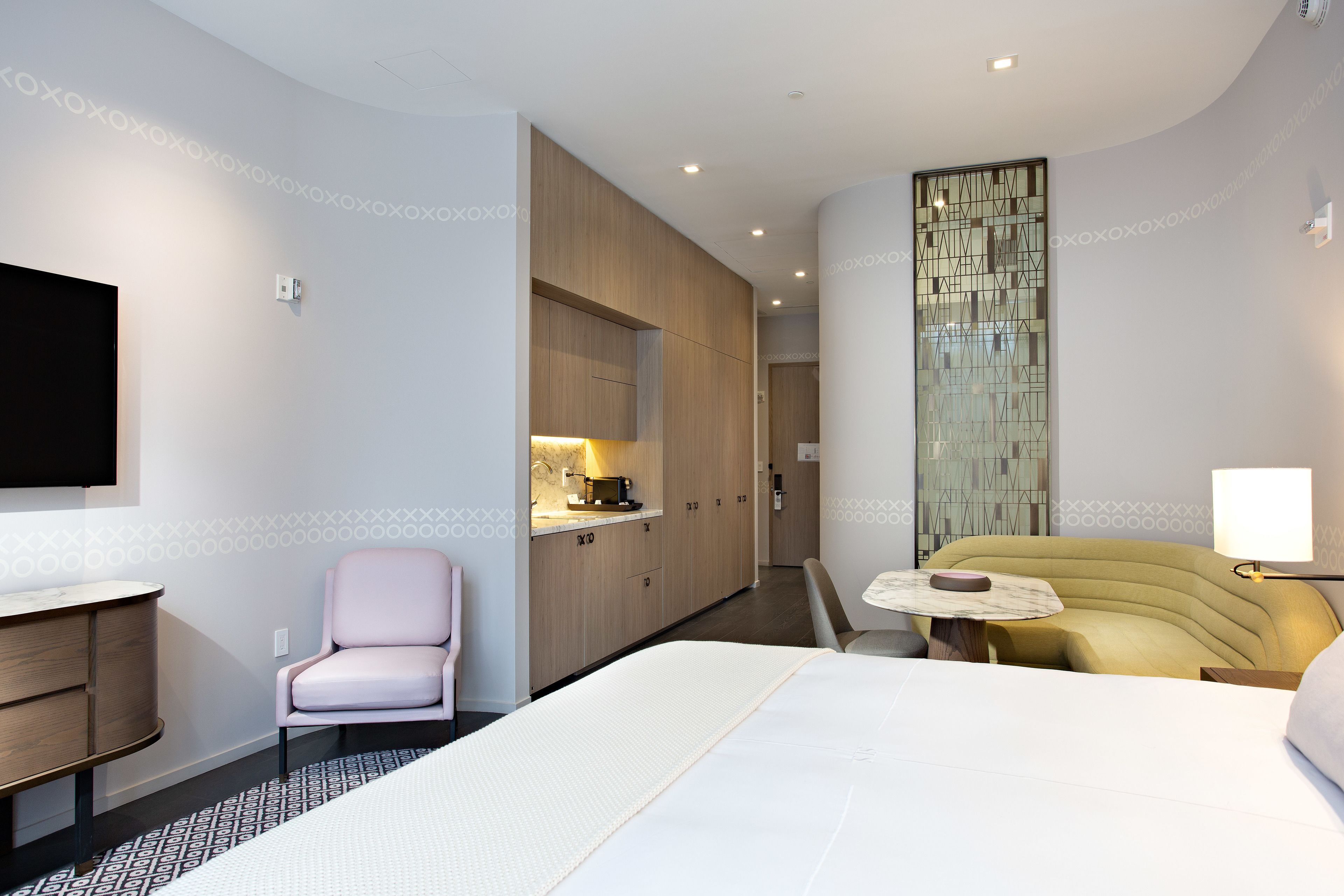 Una habitación de hotel moderna con una cama grande, un escritorio pequeño, una silla, un sofá y un televisor montado en la pared.